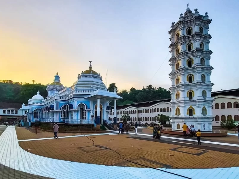 Visitors at Shree Mangesh Temple in Goa during golden hour, featuring blue domes, tall white tower and palm backdrop, perfect spiritual experience with Goa tour package. 