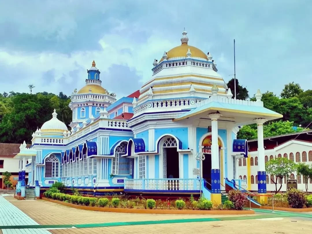 Shree Mangesh Temple in Goa during cloudy day, featuring blue domes, golden spires, white pillars and green hill backdrop, perfect spiritual experience with Goa tour package.