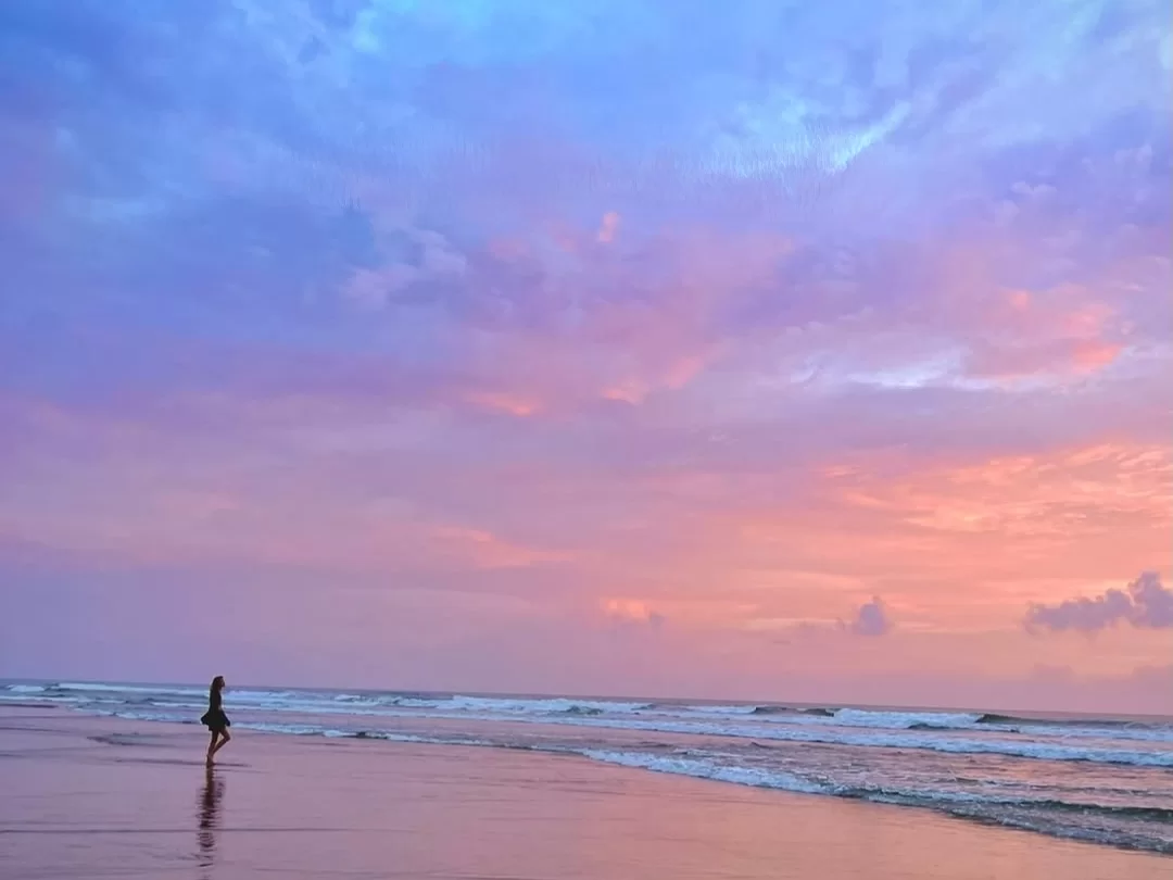 Woman strolling at Arambol Beach Goa during vibrant sunset, featuring pink-purple skies, gentle waves, wet sand reflections, perfect romantic experience with Arambol tour package.