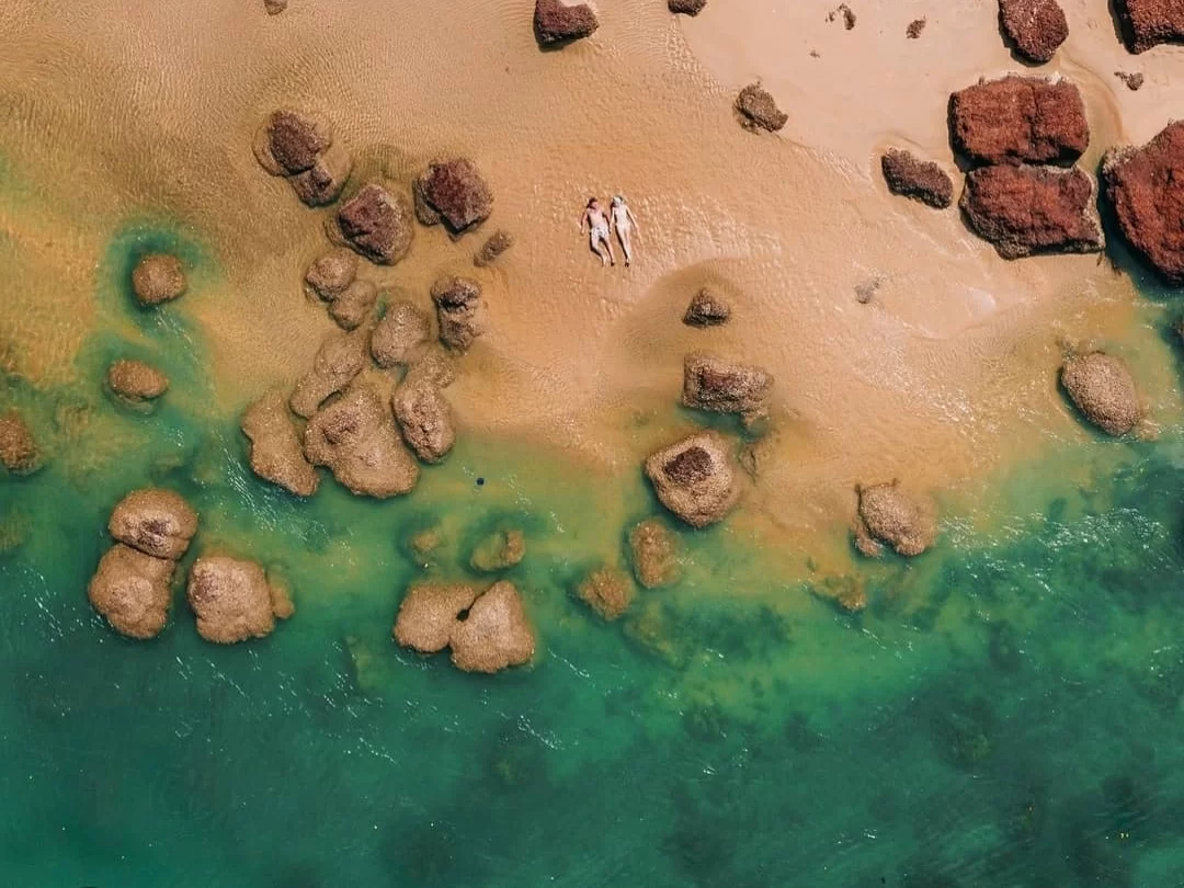 Couple relaxing at Arambol Beach Goa during sunny day, featuring red granite boulders, turquoise waters, sandy shore, perfect romantic experience with Arambol tour package.