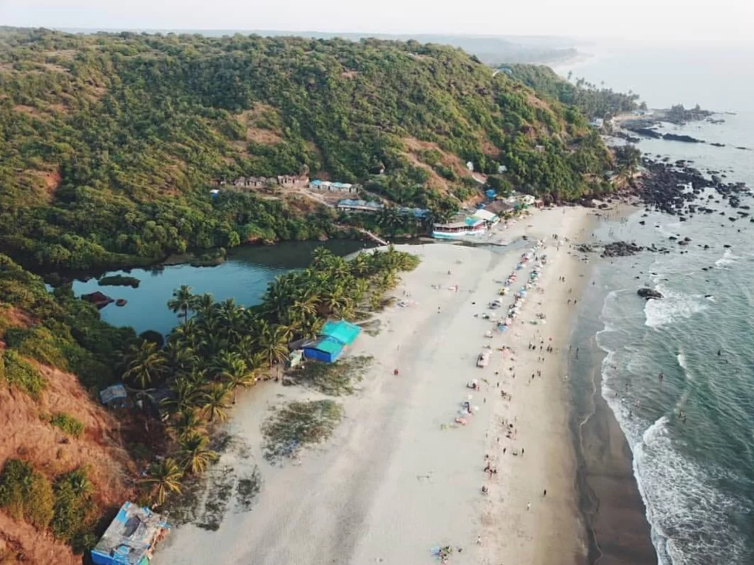 Aerial view of Arambol Beach Goa during misty morning, featuring Sweet Water Lake, green hills, beach shacks, crowds, waves, perfect bohemian experience with Arambol tour package. 