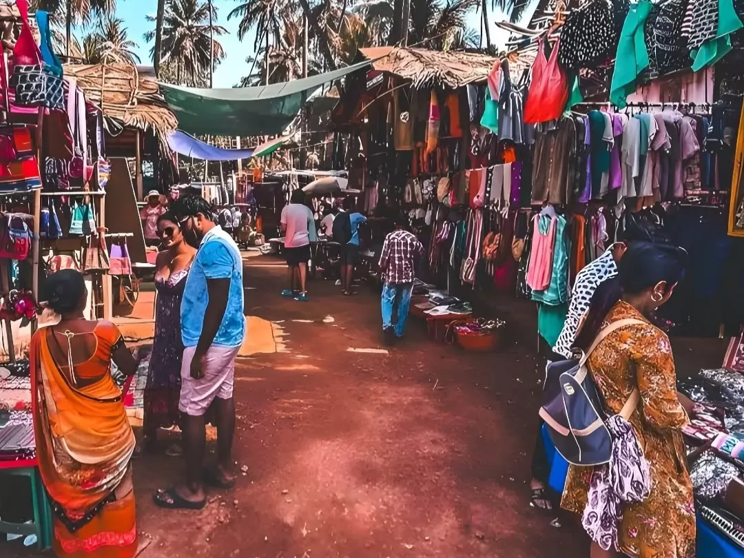 Shoppers browsing colorful clothes at Anjuna Flea Market in Goa during sunny day, featuring thatched stalls palm trees, perfect shopping adventure experience with Goa beach tour package.