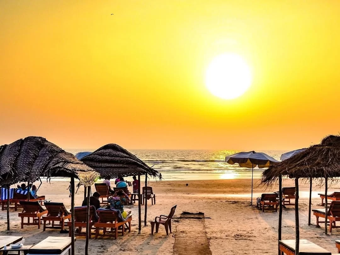 Ashvem Beach North Goa at golden sunset, featuring thatched beach umbrellas, wooden loungers, people relaxing, sandy shore, Arabian Sea waves, perfect romantic beach experience with North Goa sunset beach tour package.