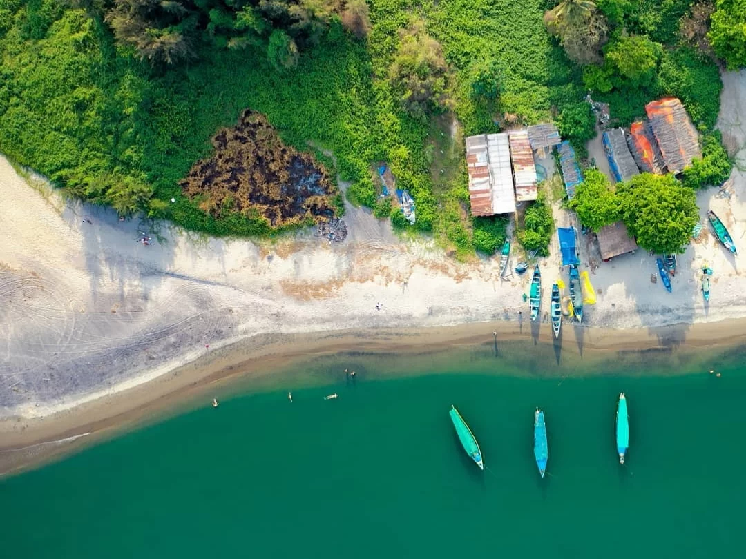 Drone aerial view of Mandrem Beach North Goa fishing village, featuring colorful traditional boats anchored turquoise lagoon white sandy shore lush green mangroves huts seaweed patches, perfect coastal village experience with Mandrem Beach Goa tour packag