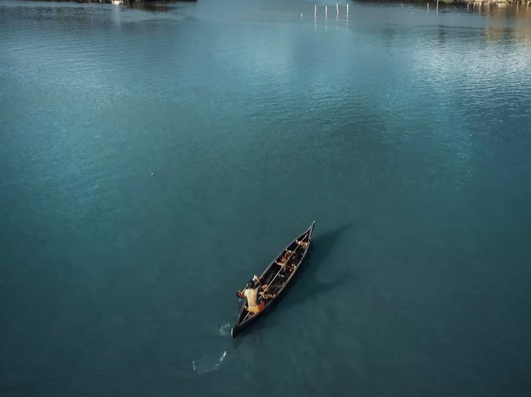 Drone aerial of traditional wooden country boat with rowers sailing on Vembanad Lake Kerala during bright day, featuring turquoise waters, distant shores, perfect local fishing life experience with Alleppey backwater village tour package.