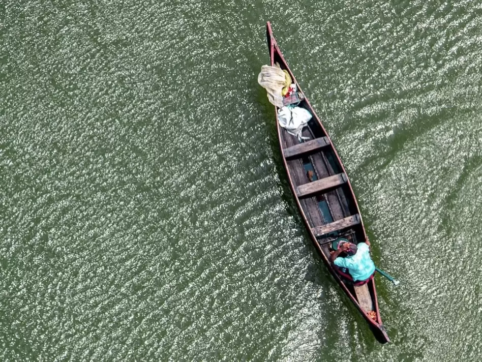 Drone aerial of traditional wooden chur boat with fisherman and nets on Vembanad Lake Kerala during sunny day, featuring turquoise ripples, distant palms, perfect local fishing life experience with Alleppey backwater village tour package. 