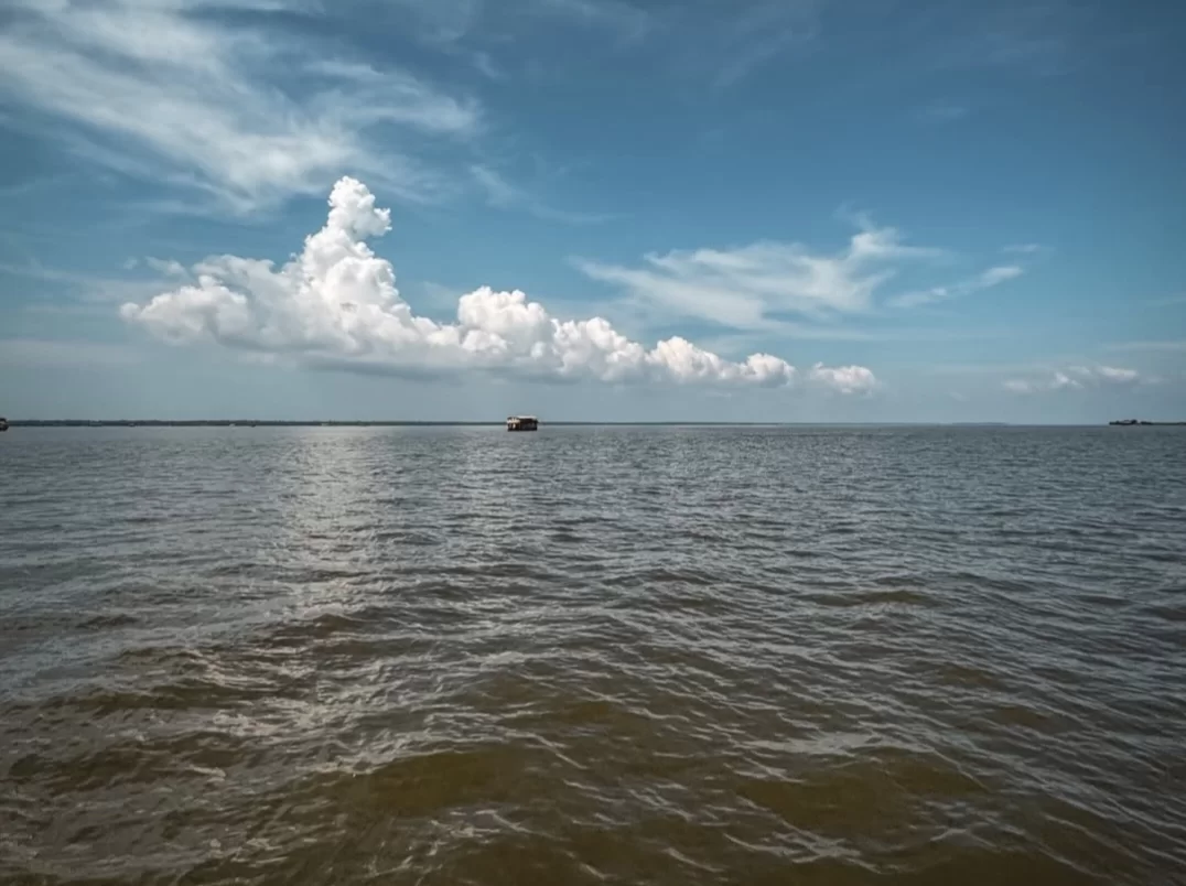 Vast Vembanad Lake Kerala under partly cloudy blue sky, featuring distant houseboat, calm reflective waters, expansive horizon, perfect serene landscape experience with Alleppey backwater houseboat cruise tour package. 
