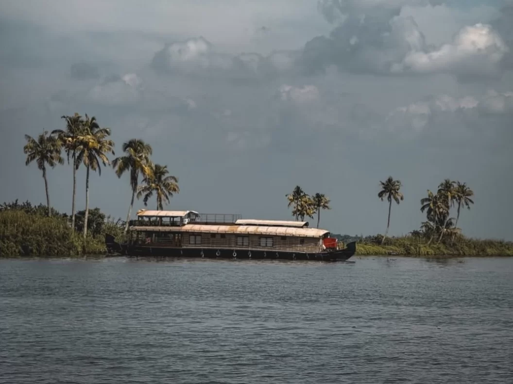 Luxury houseboat anchored on Vembanad Lake Kerala amid palm-fringed banks under overcast sky, featuring multi-deck wooden vessel, lush greenery, calm waters, perfect romantic getaway experience with Alleppey overnight houseboat cruise tour package.