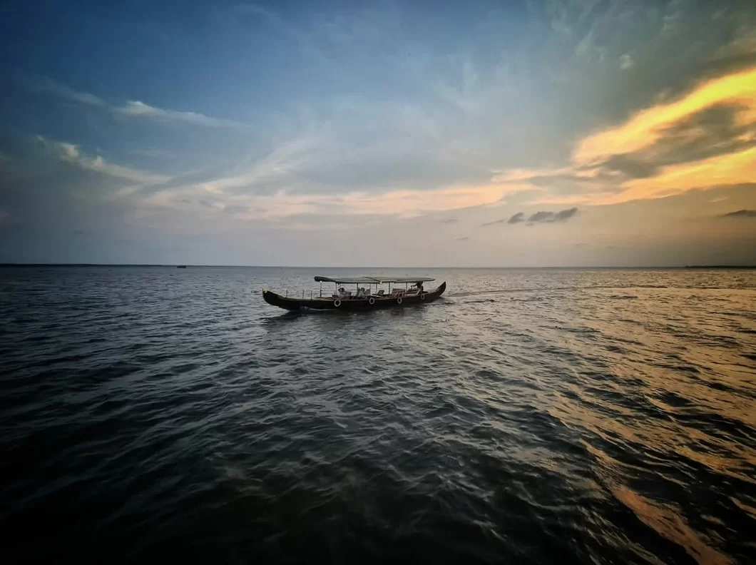 Houseboat cruising on Vembanad Lake Kerala at sunset, featuring traditional wooden vessel with passengers, orange golden sky, wispy clouds, expansive calm waters, perfect romantic luxury experience with Alleppey houseboat overnight tour package. 
