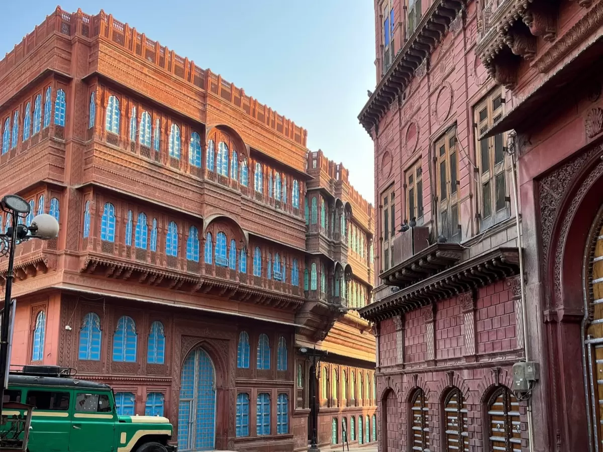 Rampuriya Haveli Bikaner Rajasthan grand sandstone facades, featuring multi-story red buildings blue windows jharokhas vintage jeep foreground, perfect Rajasthan tour packages.