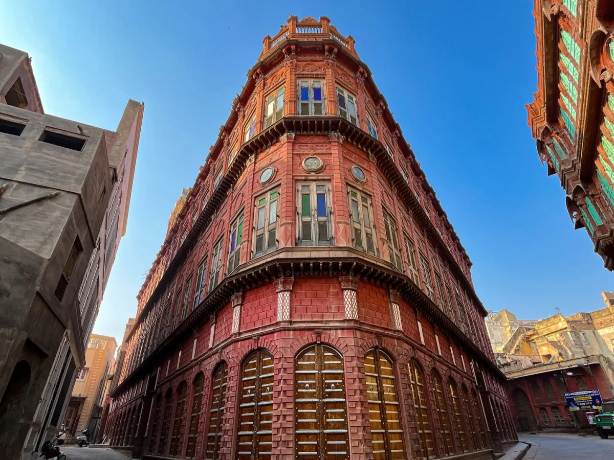 Rampuriya Haveli Bikaner Rajasthan panoramic corner view, featuring tall red sandstone structure arched doors colorful windows adjacent buildings blue sky, perfect Rajasthan tour packages.