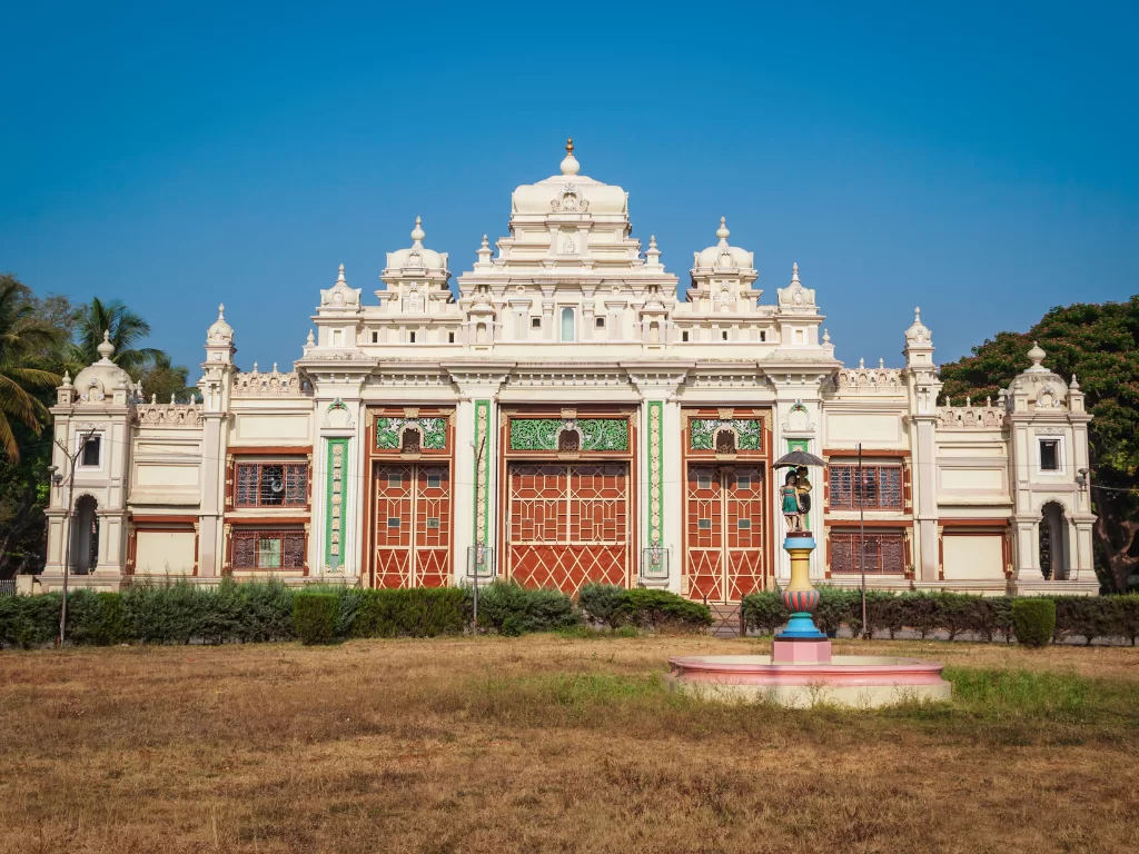 Jaganmohan Palace in Mysore during sunny daytime, featuring symmetrical ivory facade, green accents and foreground statue, perfect heritage Karnataka tour package