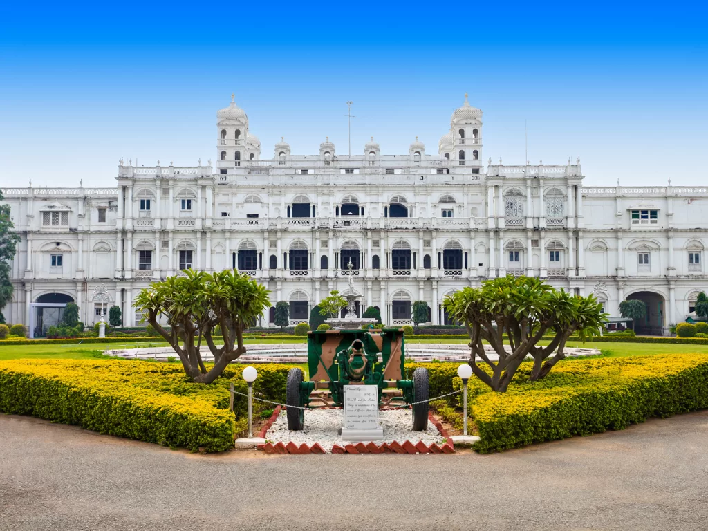 Jai Vilas Palace Gwalior during clear afternoon, featuring white Italianate facade with domed towers before manicured gardens showcasing silver royal carriage display amid yellow flower borders, perfect heritage experience in Madhya Pradesh tour package.