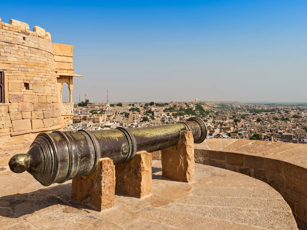 Ancient cannon at Jaisalmer Fort during clear day, featuring sandstone walls & city skyline views, perfect adventure experience Rajasthan tour packages.