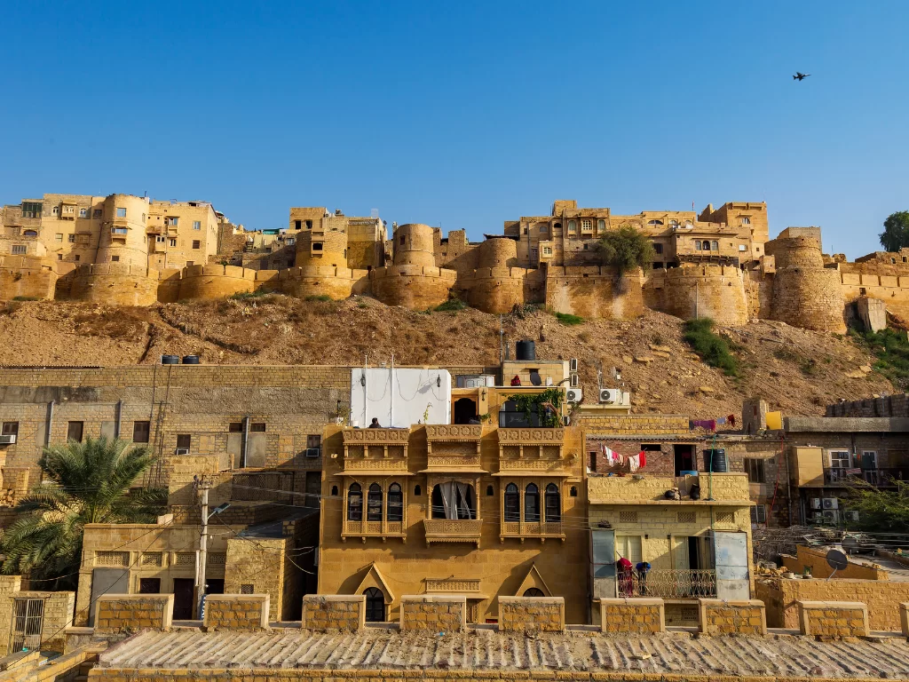Golden havelis below Jaisalmer Fort during blue sky, featuring ramparts & palms, perfect adventure experience Rajasthan tour packages.