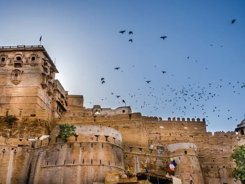 Majestic bastions at Jaisalmer Fort during clear blue sky, featuring flying birds & flags, perfect adventure experience Rajasthan tour packages.