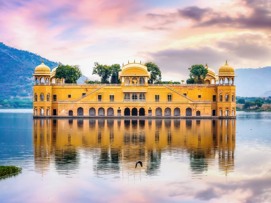 Jal Mahal palace at Jaipur during pink sunset clouds, featuring stunning lake reflections trees domes Aravalli hills, perfect romantic experience Rajasthan tour packages.
