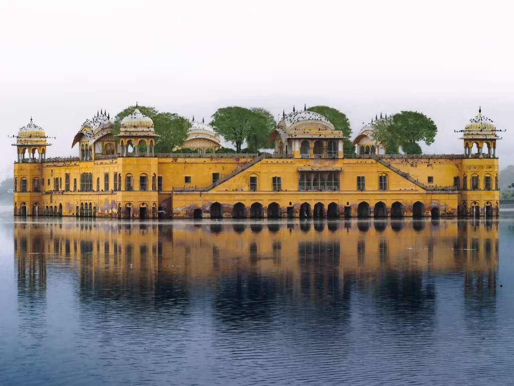 Jal Mahal palace at Jaipur during misty morning, featuring stunning lake reflections lush trees domes Aravalli hills, perfect romantic experience Rajasthan tour packages.