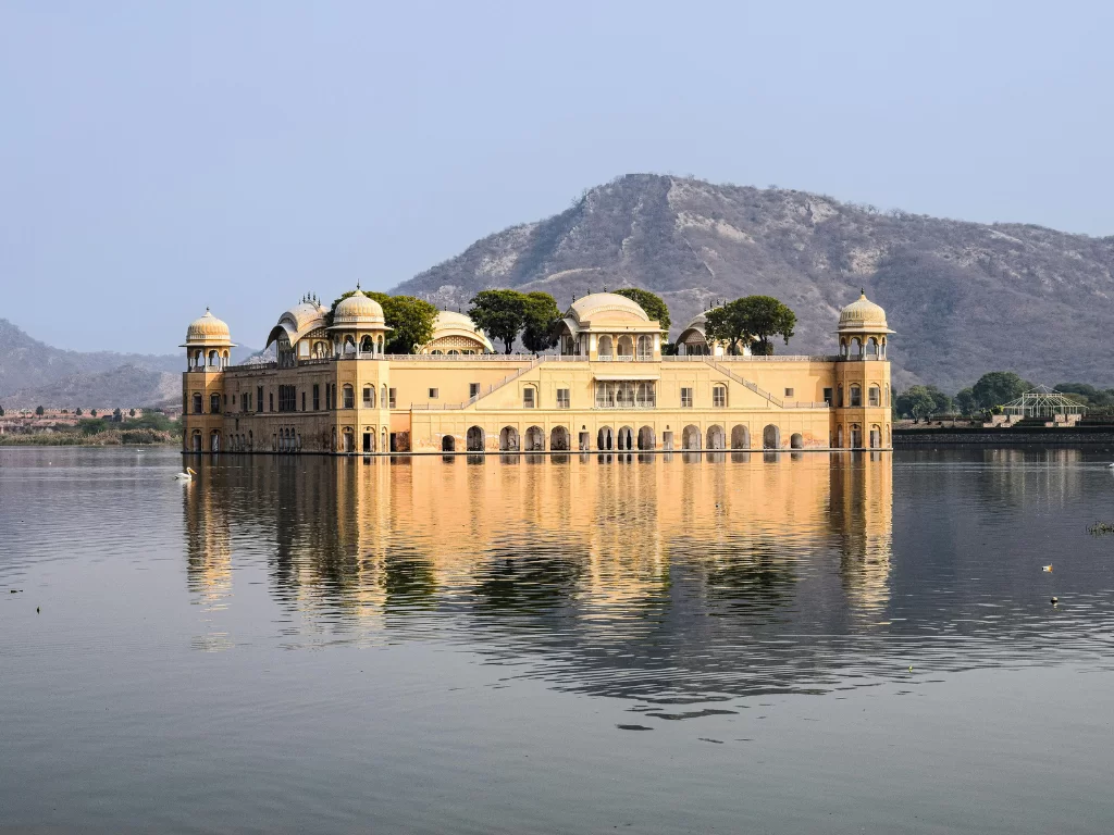 Jal Mahal palace at Jaipur during clear daytime, featuring perfect lake reflections trees domes Aravalli mountains, perfect romantic experience Rajasthan tour packages.