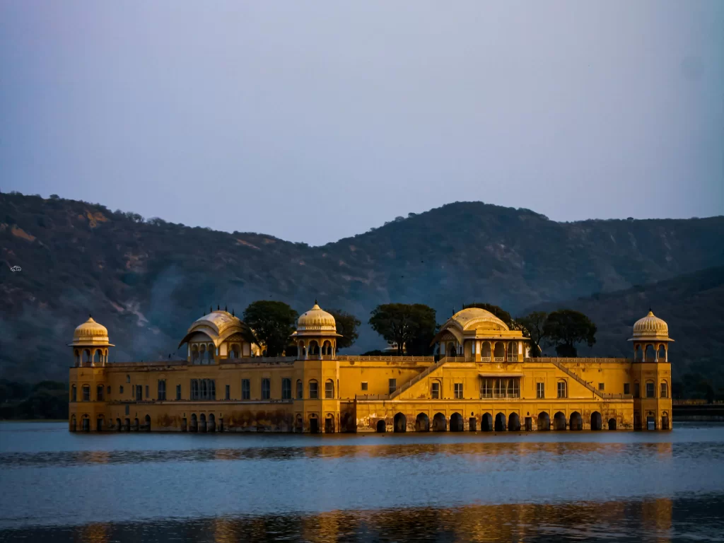 Jal Mahal palace at Jaipur during twilight dusk, featuring lake reflections trees domes Aravalli mountains, perfect romantic experience Rajasthan tour packages.