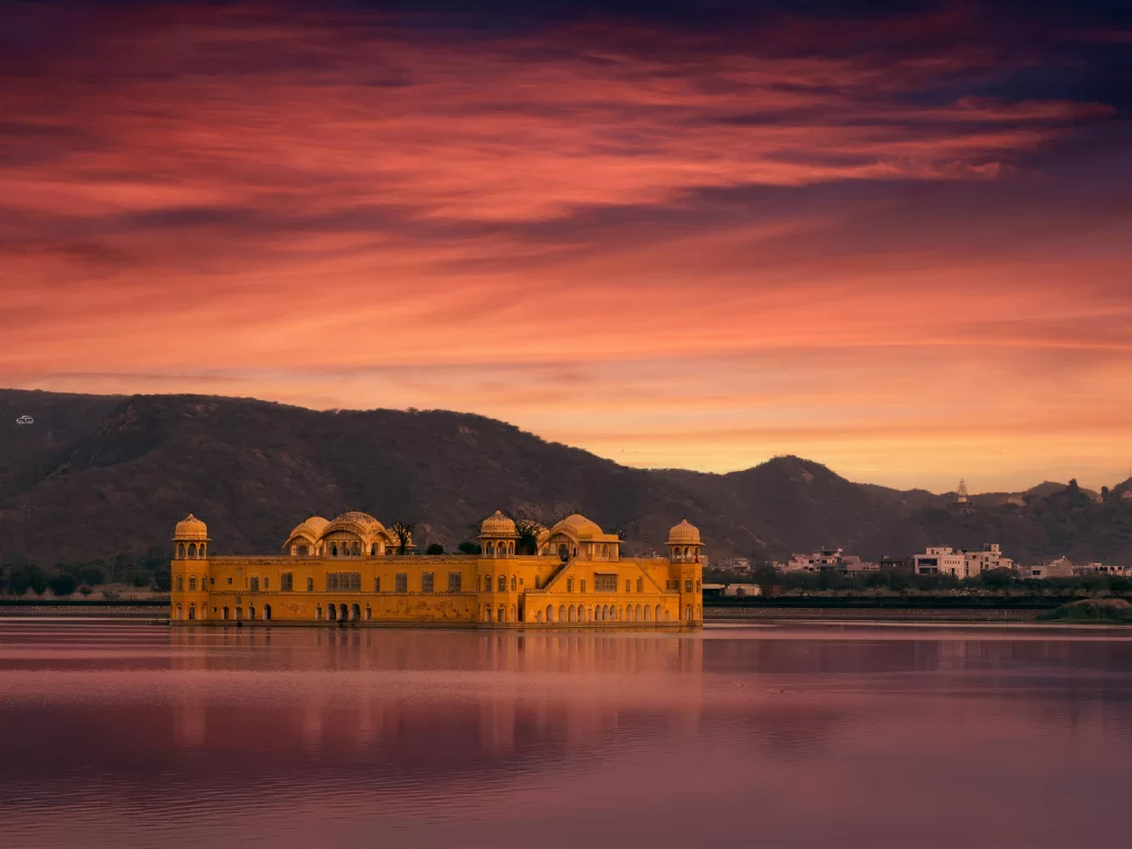 Jal Mahal at Jaipur during pink sunset glow, featuring lake reflections Aravalli mountains domes, perfect romantic experience Rajasthan tour packages.