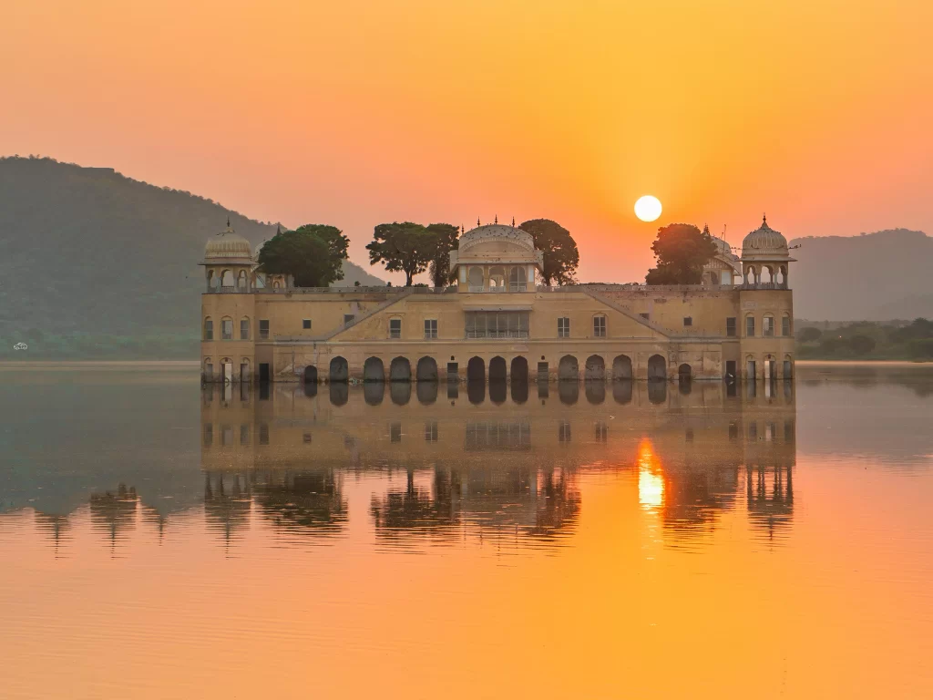Jal Mahal palace at Jaipur during vibrant sunset, featuring lake reflections Aravalli hills trees, perfect romantic experience Rajasthan tour packages.