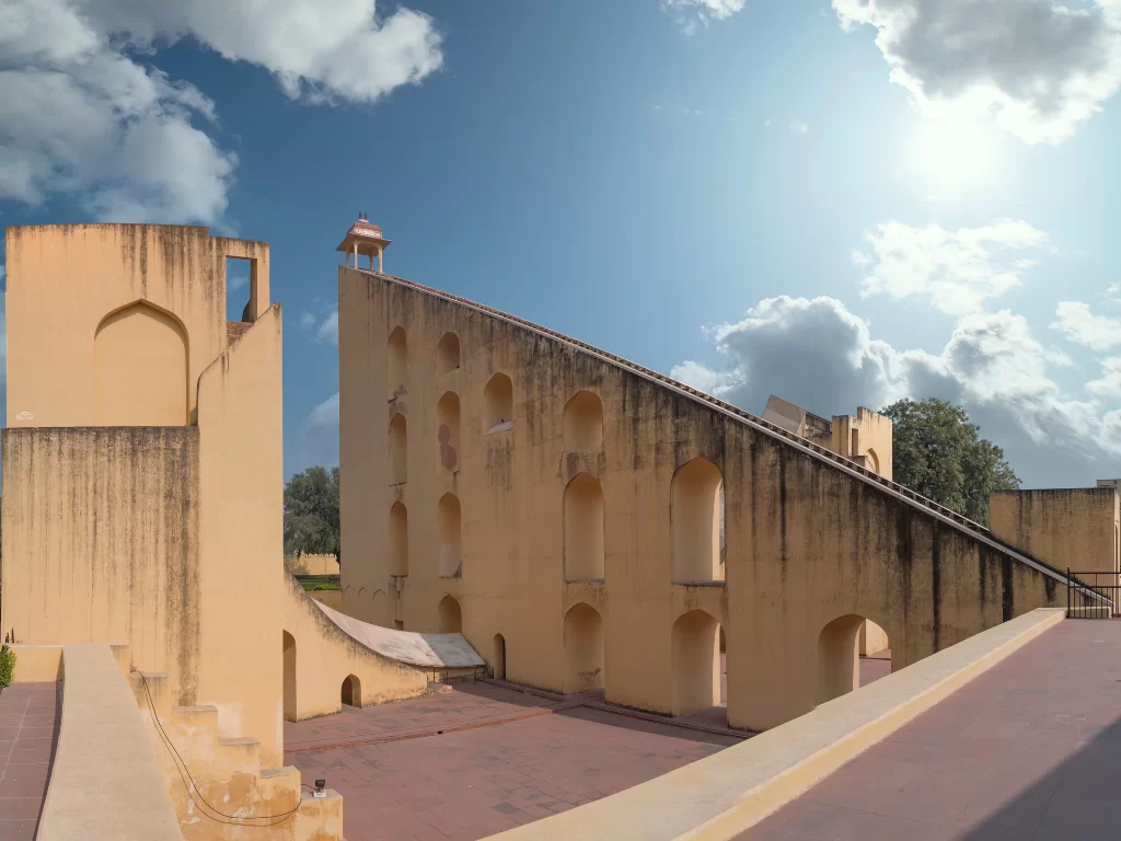 Jantar Mantar at Jaipur during sunny partly cloudy day, featuring tall astronomical instruments arches dome trees, perfect heritage experience Rajasthan tour packages.