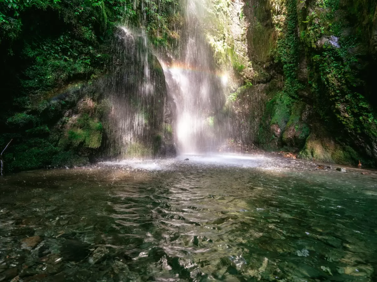 Jibhi Waterfall Tirthan Valley Himachal Pradesh during sunny conditions with rainbow spray, featuring multi-tier cascading falls turquoise pool lush green forested mossy rocky gorge backdrop, perfect cultural experience Himachal tour package.