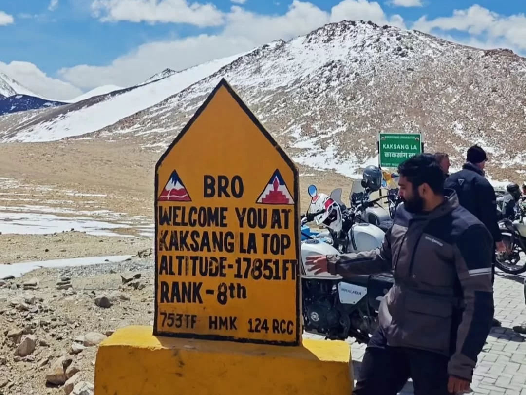 Motorcyclists at BRO sign Welcome You at Kaksang La Top Altitude 17851 FT Rank-8th during partly cloudy skies, featuring snowy hills, perfect biking adventure Ladakh tour package.