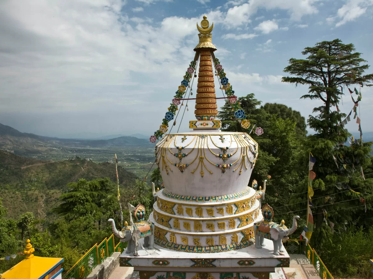 Kalachakra Temple complex McLeod Ganj Dharamshala Himachal Pradesh during partly cloudy skies, featuring ornate white multi-tier Tibetan stupa golden spire prayer flags deodar pine forested mountain valley backdrop, perfect cultural experience Himachal to