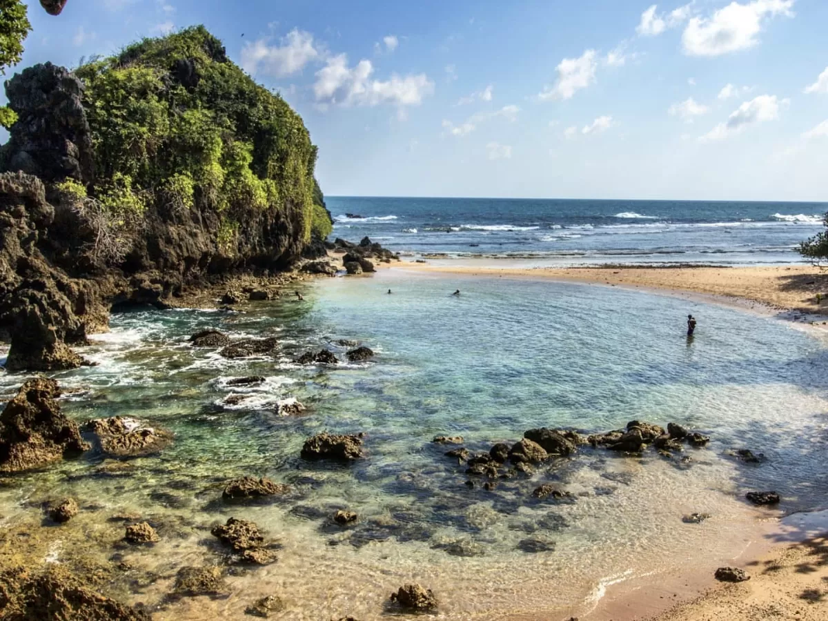 Lush green cliff rocky outcrop turquoise natural pool white sandy beach ocean waves distant swimmer partly cloudy blue sky Kalapathar Beach Havelock Island Andaman Islands, perfect pristine sunrise beach eco-tourism Andaman package.