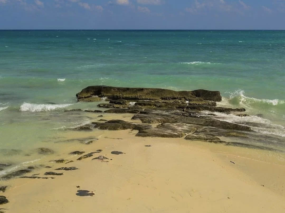 Sandy beach meeting turquoise ocean waves cluster black volcanic rocks breaking surf partly cloudy blue sky Kalapathar Beach Havelock Andaman, signature black pebble shoreline eco-tourism Andaman Islands package.