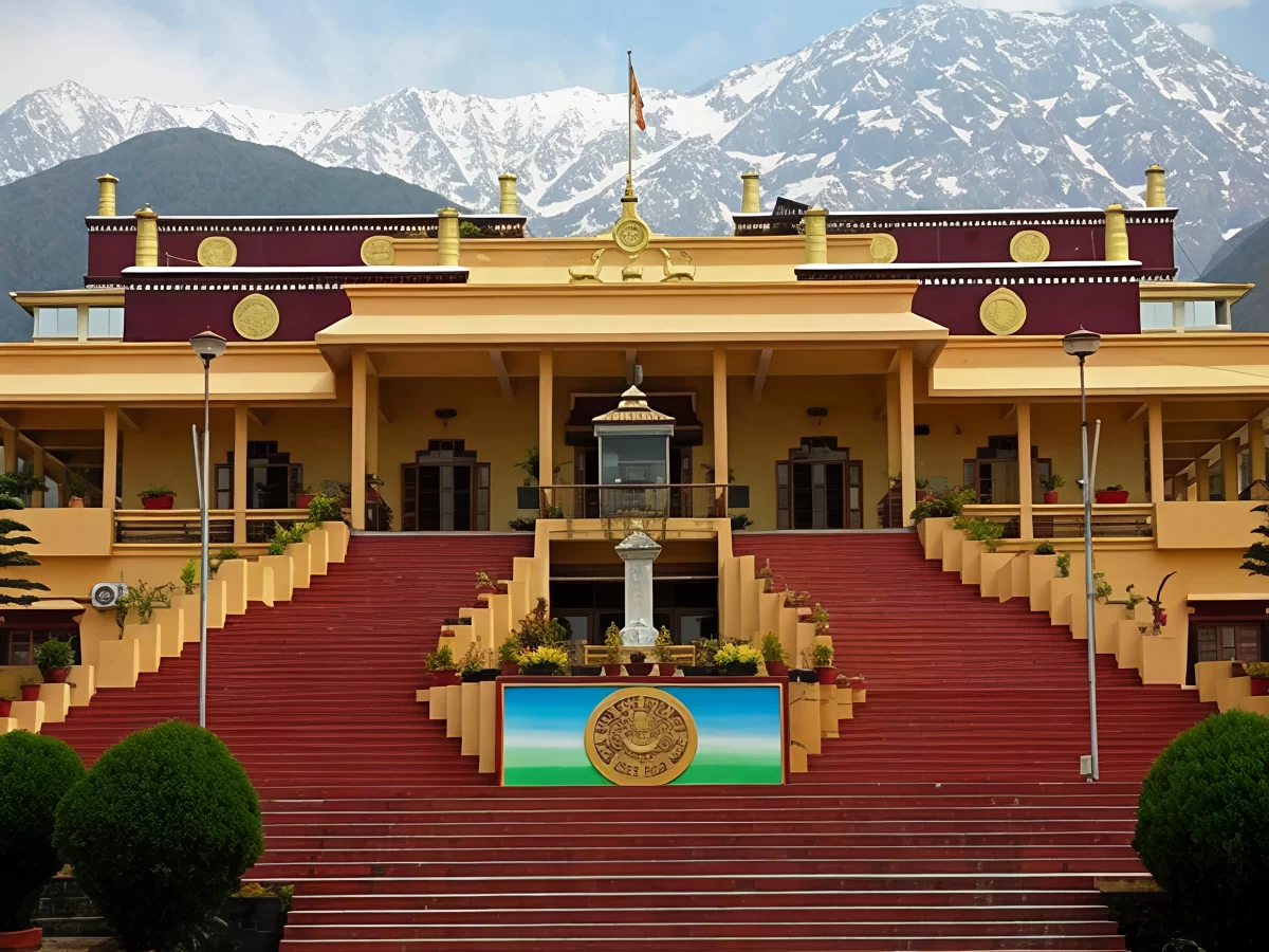 Kangra Art Museum in Dharamshala featuring a grand yellow and maroon building with wide red staircases, decorative emblem at the entrance, and snow-capped Dhauladhar mountains in the background.