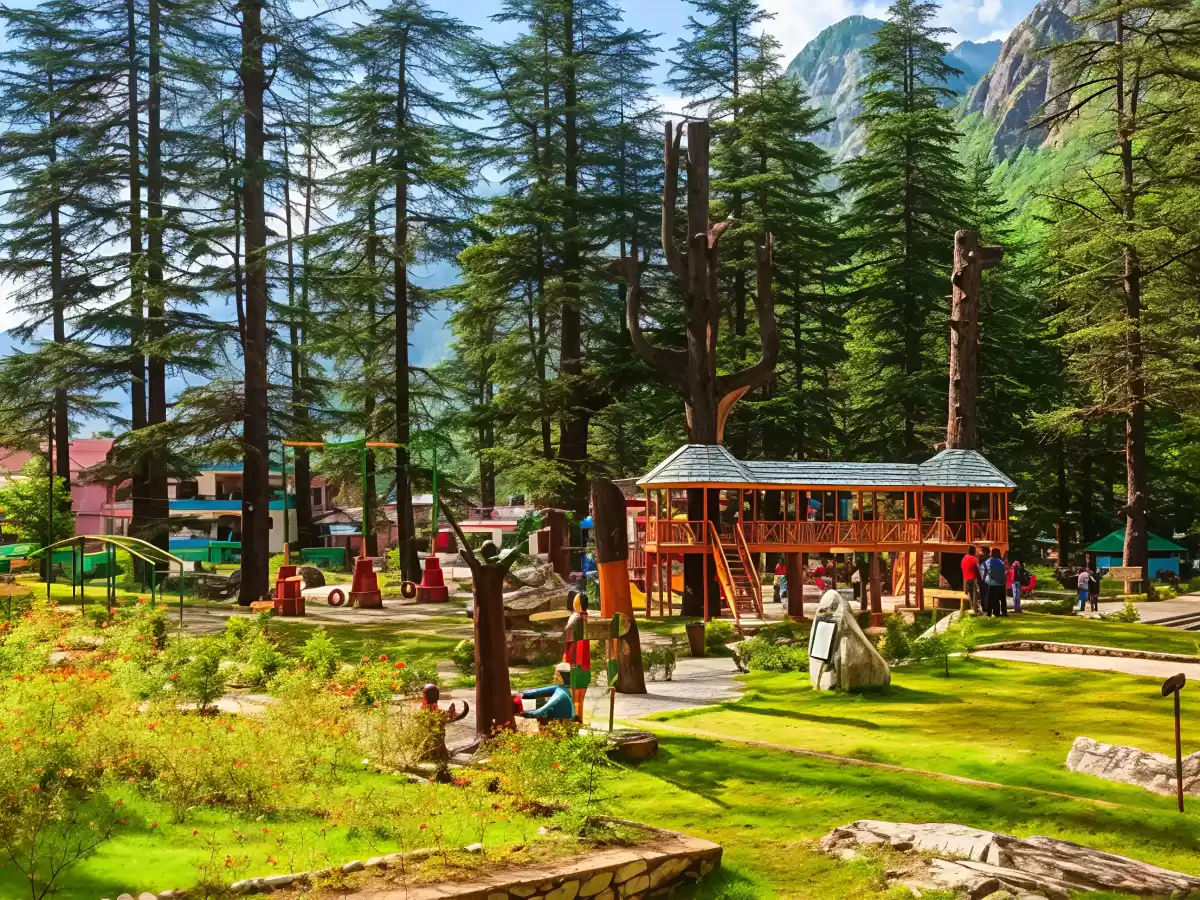 Scenic view of Nature Park in Kasol featuring tall pine trees, a wooden treehouse-style structure, green lawns, walking paths, and mountains in the background with visitors exploring the park.