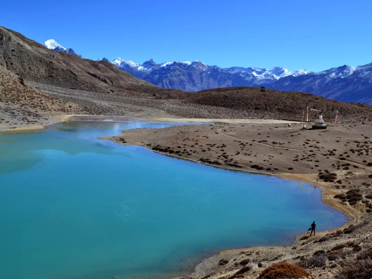 Dhankar Lake near Kibber Village Kaza during clear skies, featuring turquoise water stupa hiker rocky shore snow mountains, perfect adventure experience Himachal tour package.