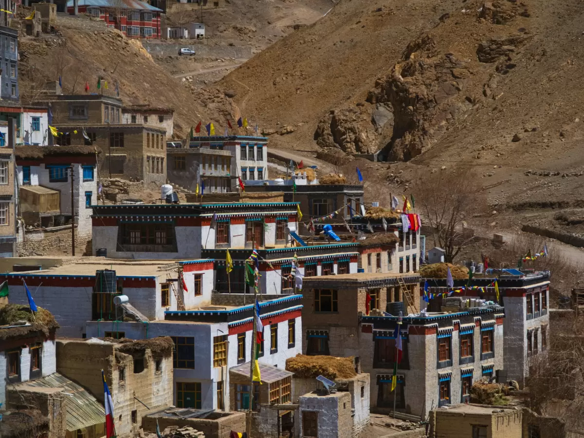 Kibber Village near Kaza during sunny skies, featuring white multi-story houses colorful prayer flags rocky hills, perfect cultural experience Himachal tour package.