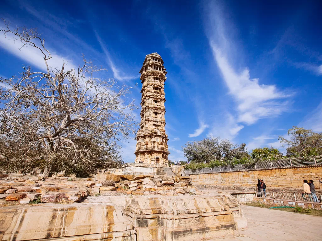 Close-up of Kirti Stambh tower at Chittorgarh Fort in Rajasthan under clear blue sky, featuring multi-tiered architecture and visitors, perfect Rajasthan tour package.