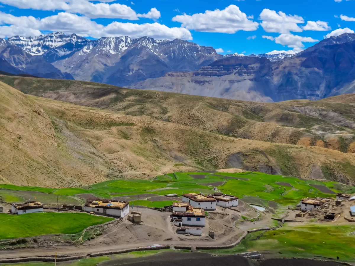 Komic Village near Kaza during partly cloudy skies, featuring whitewashed houses green fields brown hills snow mountains, perfect cultural experience Himachal tour package.