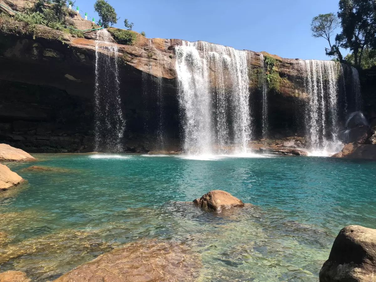 Krang Shuri Falls Cherrapunji during sunny day, featuring cave waterfall turquoise pool rocks trees blue sky foreground vegetation, perfect adventure nature Cherrapunji Meghalaya tour package.