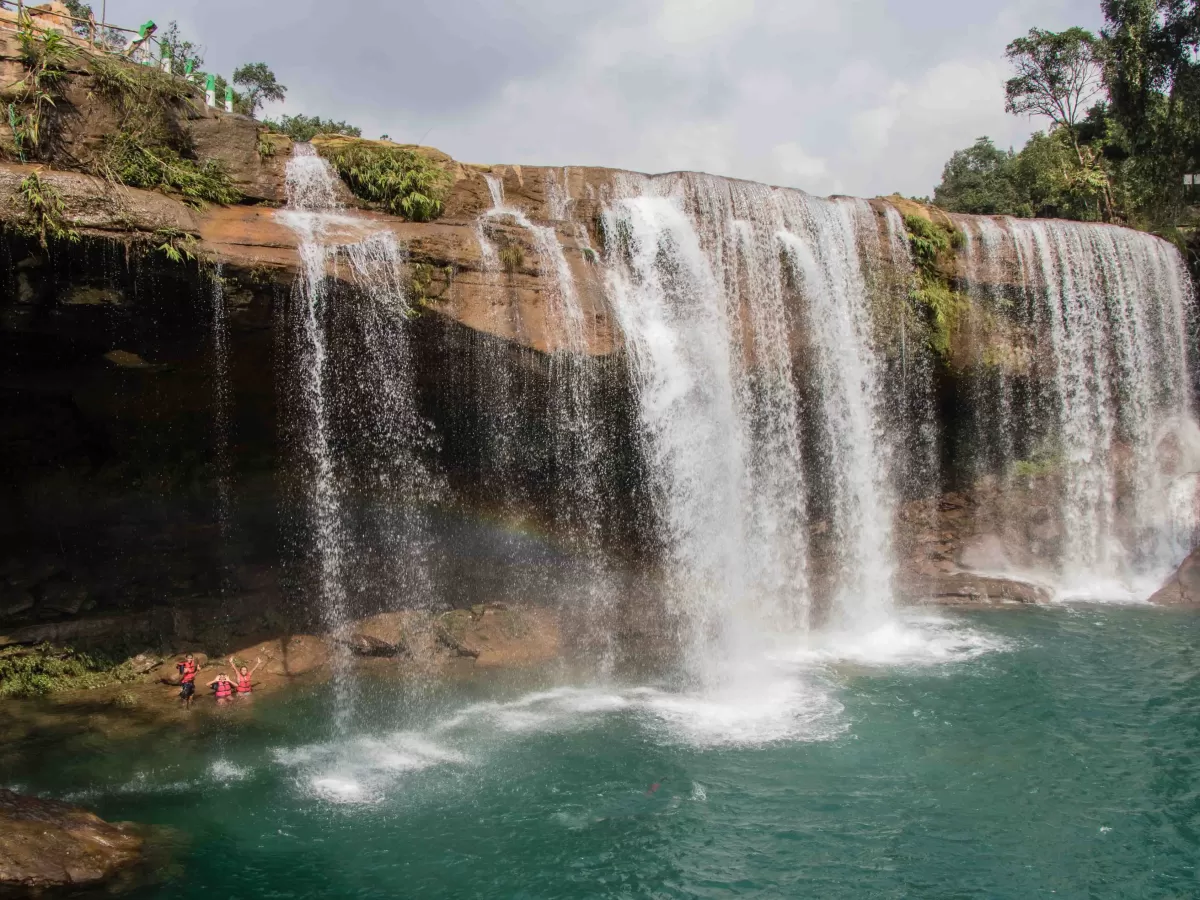 Krang Shuri Falls Cherrapunji during cloudy day, featuring multi-tiered waterfall turquoise pool swimmers viewing platform trees rocky cliffs, perfect adventure nature Cherrapunji Meghalaya tour package.