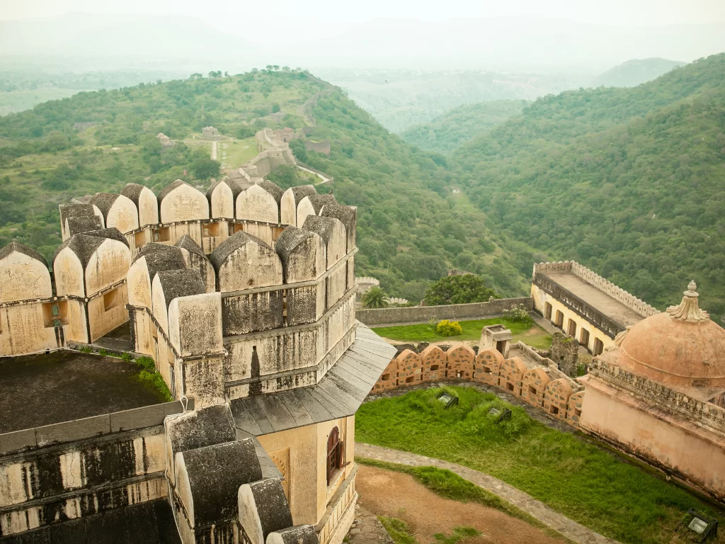 Kumbhalgarh Fort bastions during misty haze, featuring curved merlons, dome pavilion, green hills, perfect adventure experience Rajasthan tour packages.