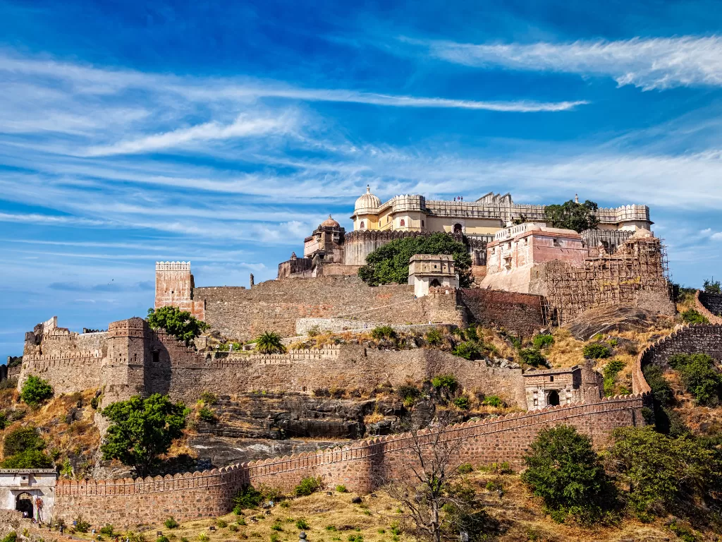 Kumbhalgarh Fort during partly cloudy sky, featuring multi-level palaces, bastions, dome tops, hillside setting, perfect adventure experience Rajasthan tour packages. 