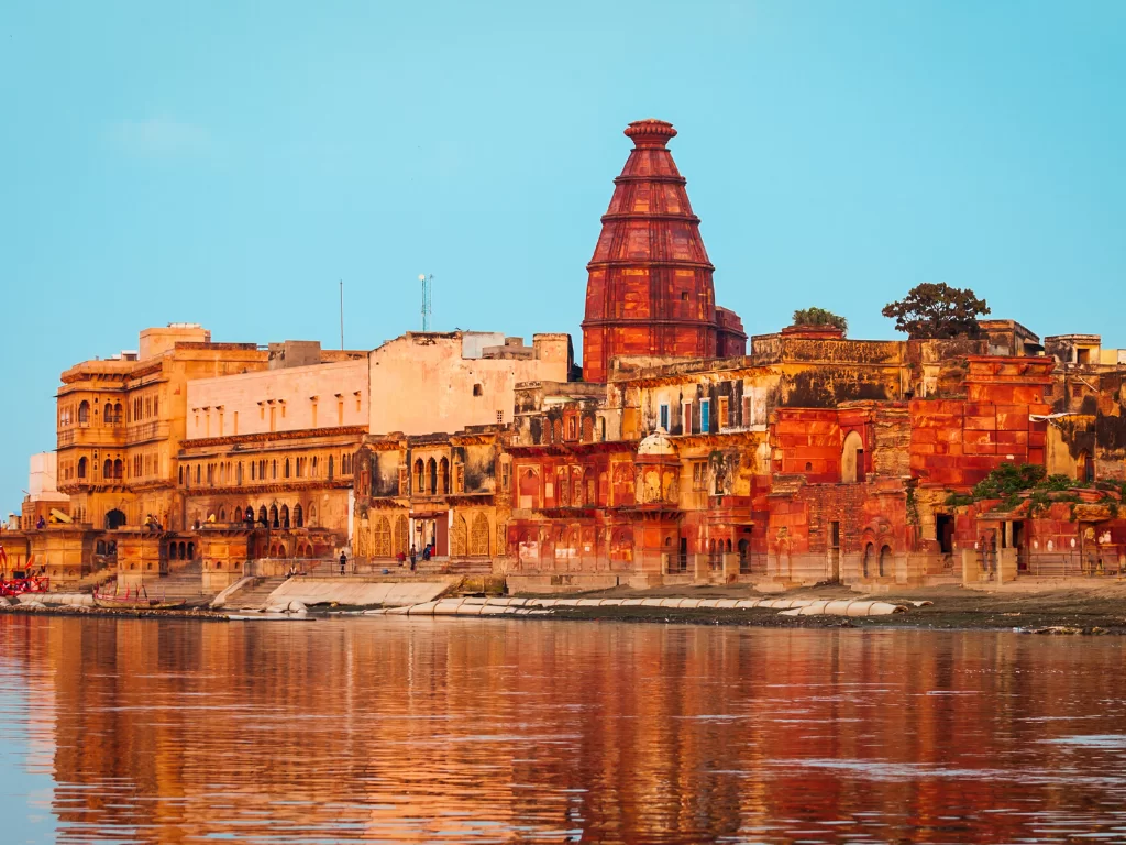 Red sandstone palaces at Kusum Sarovar during golden hour, featuring terracotta temple spire, river reflections, perfect spiritual experience with Mathura Vrindavan tour package. 
