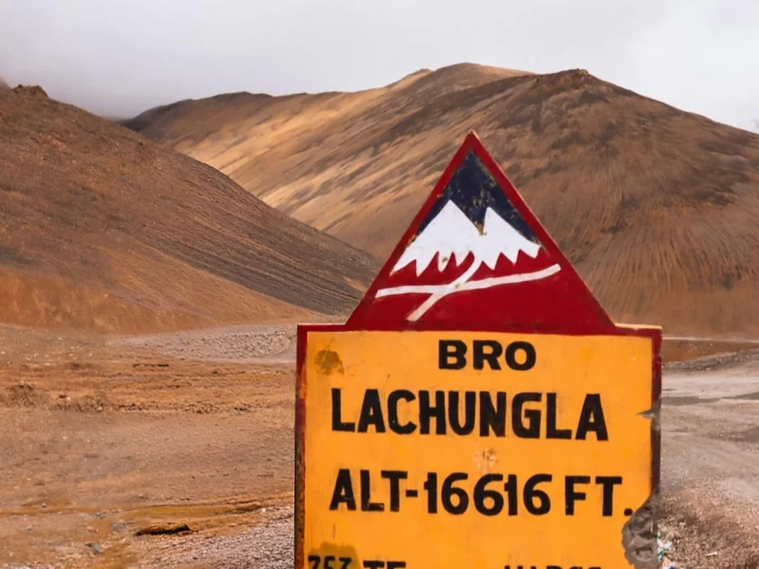 Lachulung La Pass at 16,616 ft on Leh-Manali Highway in misty weather, featuring BRO sign and rugged mountains, perfect adventure for Ladakh tour package.