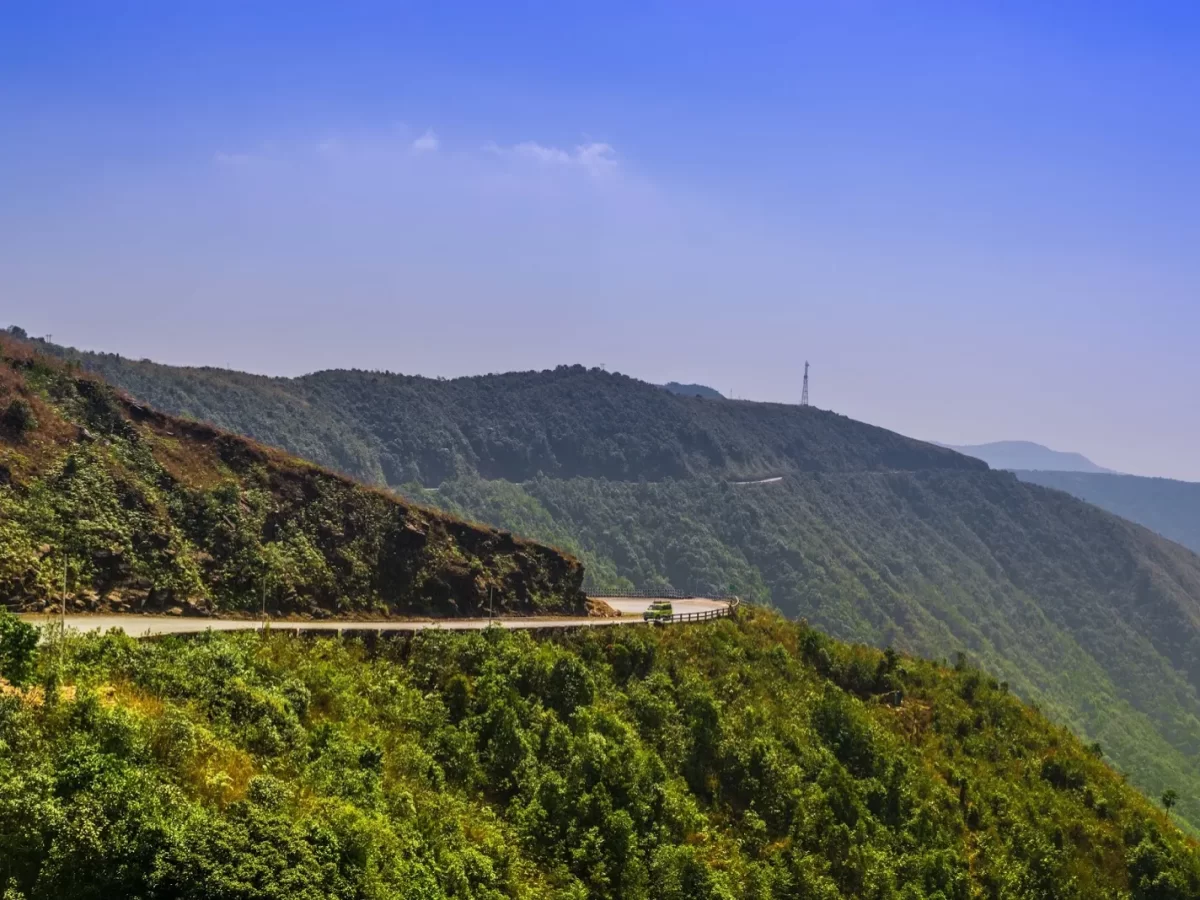 Laitlum Canyons Shillong Meghalaya winding mountain road hairpin bends lush green hills blue sky tower Shillong Peak background scenic drive viewpoint near canyons, perfect Meghalaya tour packages.