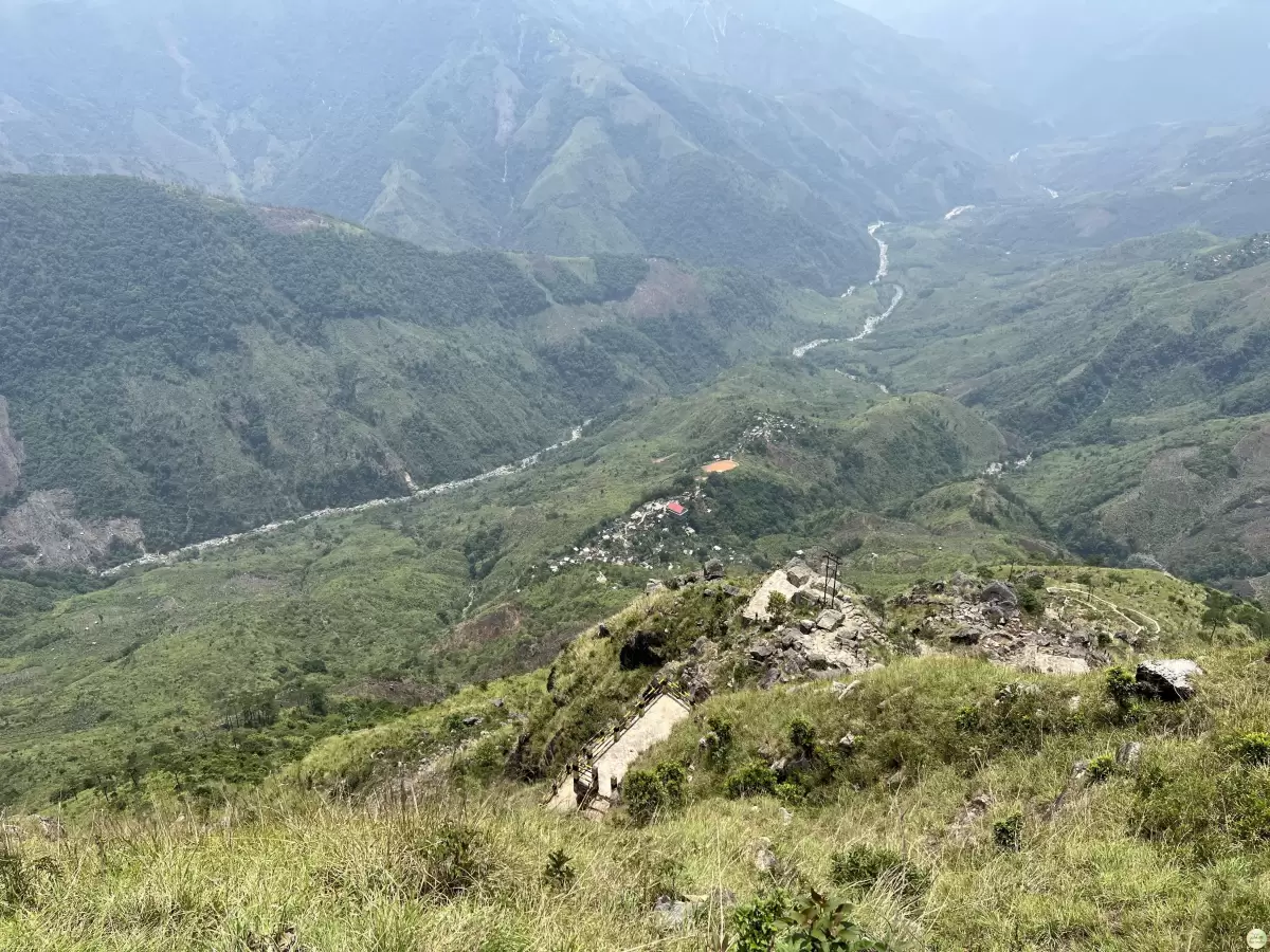 Laitlum Canyons Shillong Meghalaya panoramic valley view lush green hills winding river villages terraced fields distant blue mountains cloudy sky foreground grass rocky outcrop tiny figures viewpoint, perfect Meghalaya tour packages.