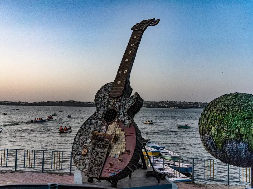 Giant guitar sculpture at Lake View in Bhopal, Madhya Pradesh, overlooking the water with boating activities in the background, a popular waterfront attraction featured in Madhya Pradesh tour packages