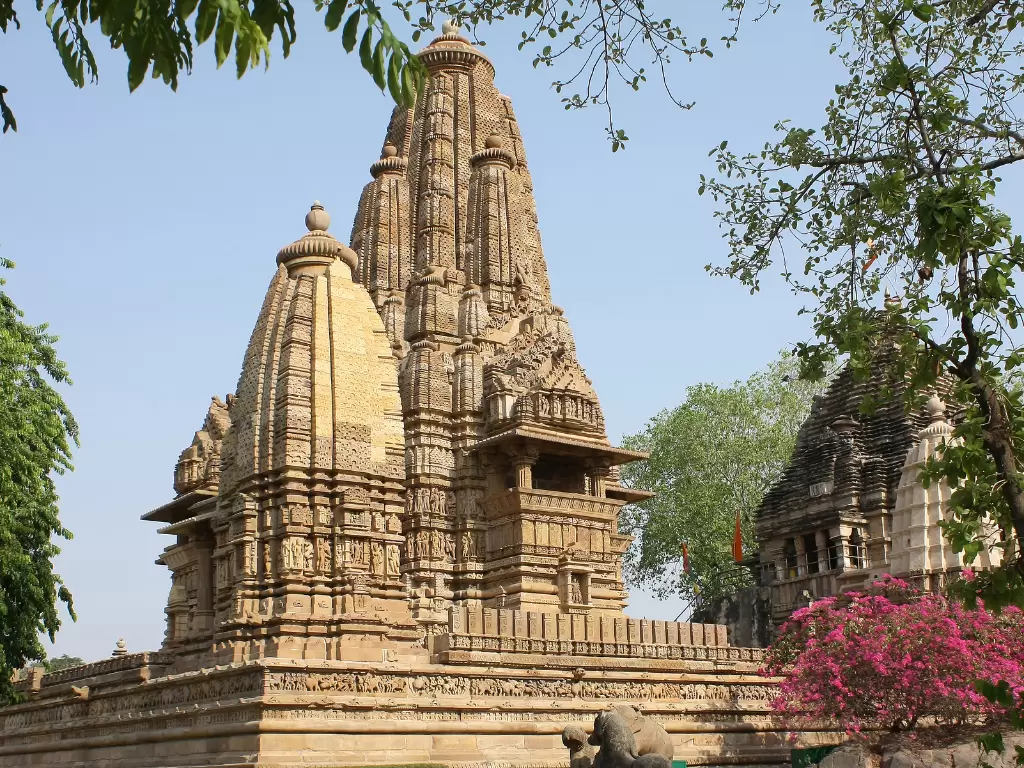 Lakshmana Temple Khajuraho tree-framed view Western Group UNESCO site, Nagara-style Vaikuntha Vishnu shrine intricate carvings adjacent shrines Chandela dynasty 930-950 AD, perfect Madhya Pradesh heritage temple tour package.