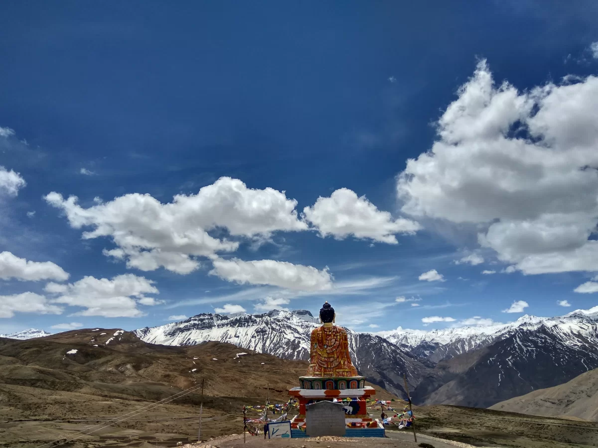Buddha statue at Langza Village near Kaza during partly cloudy skies, featuring prayer flags rocky slopes snow mountains, perfect cultural experience Himachal tour package.