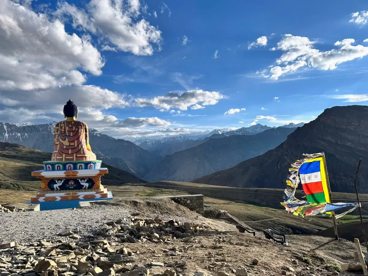 Buddha statue at Langza Village near Kaza during partly cloudy skies, featuring prayer flags rocky terrain snow mountains, perfect cultural experience Himachal tour package.